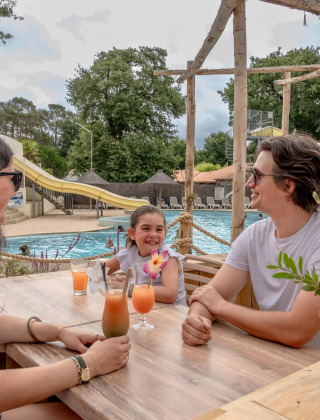 Gezin aan tafel bij het zwembad met drankjes bij Flower Camping Au Bois Des Biches in Frankrijk.