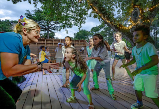 Des enfants jouent et dansent avec une adulte sur une terrasse en bois entourée d’arbres dans un parc de vacances en France.
