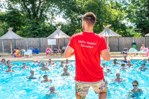 A swim instructor in a red shirt leads a water aerobics class at an outdoor pool on a sunny day.
