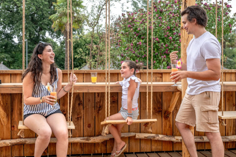 Gezin geniet van drankjes op houten terras met schommels bij Flower Camping Au Bois Des Biches.