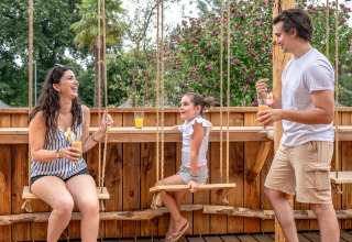 Familia disfrutando de bebidas en una terraza de madera con columpios en Flower Camping Au Bois Des Biches.