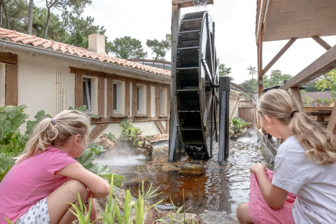 Two girls watch a large water wheel by a pond at Flower Camping Au Bois Des Biches, Pays de la Loire, France.
