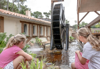 Two girls watch a large water wheel by a pond at Flower Camping Au Bois Des Biches, Pays de la Loire, France.