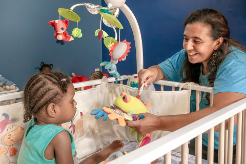 Eine Frau spielt mit einem Kleinkind im Gitterbett, umgeben von buntem Babyspielzeug und Mobiles.
