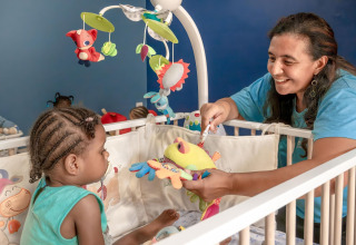 Eine Frau spielt mit einem Kleinkind im Gitterbett, umgeben von buntem Babyspielzeug und Mobiles.
