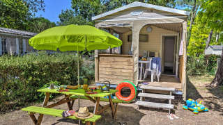 Buitenzicht op een Cabane Nature safaritent met picknicktafel, groene parasol en kleurrijk speelgoed.