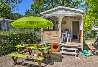 Vista esterna di una tenda safari Cabane Nature con tavolo da picnic, ombrellone verde e giochi colorati.