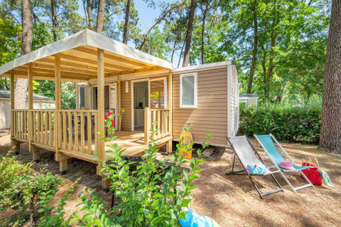 Stacaravan Confort chalet i en skov, med træterrasse, liggestole og udendørs ferieindretning.
