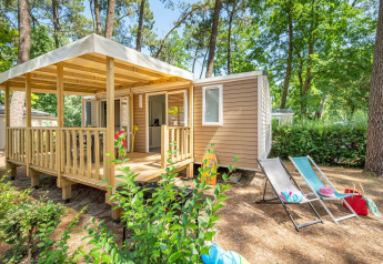 Stacaravan Confort chalet in het bos, met houten terras, ligstoelen en vakantie-attributen.