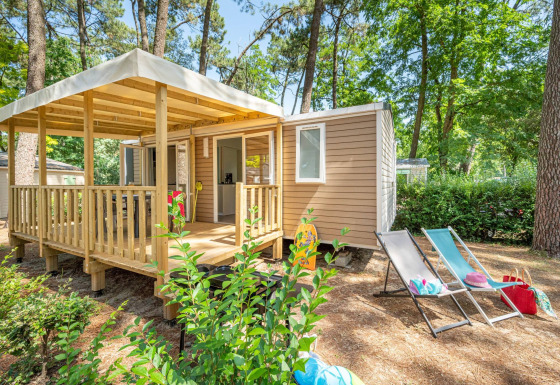 Stacaravan Confort chalet i en skov, med træterrasse, liggestole og udendørs ferieindretning.
