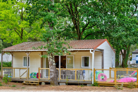 Cottage Family chalet met houten terras, tussen groene bomen en kleurrijk buitenspeelgoed.