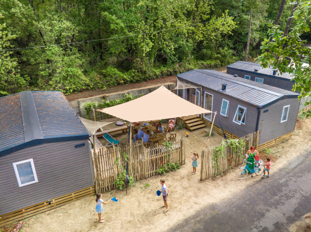 Luchtfoto van glamping-accommodatie met moderne huisjes, families genieten buiten onder een zonnedoek.