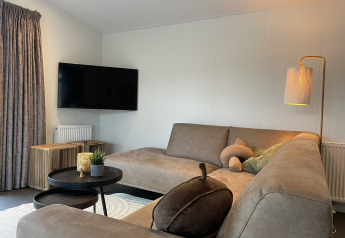 Salón moderno en una cabaña con sofá gris, televisor, lámpara de pie y mesas de madera en De Witte Berg.