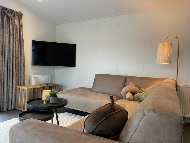 Moderne woonkamer in een lodge met grijze zetel, tv, vloerlamp en houten tafels bij De Witte Berg.