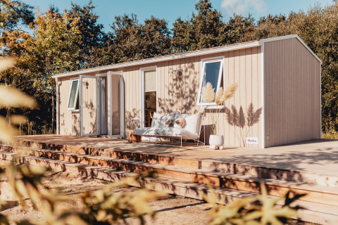 Moderne glampinghut met houten terras, buitensofa en planten, omgeven door natuur in het bos.
