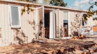 Udsigt til et glamping-hus med åbne døre, terrasse og hyggelig siddeplads i solrigt vejr.