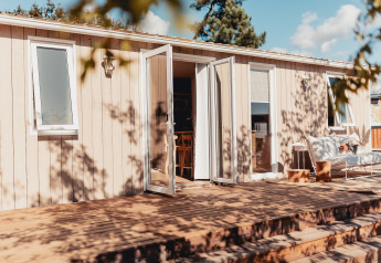 Alloggio glamping con porte aperte, terrazza in legno e zona relax sotto il sole estivo.