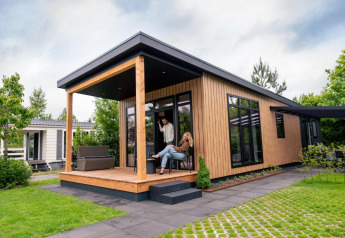 To personer slapper af på verandaen af en moderne Sunlodge-lodge omringet af grøn have og træer.
