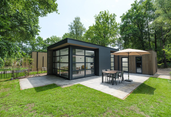Moderne Bosvilla Comfort lodge met ruime ramen, terras, parasol en eettafel in een groene omgeving.