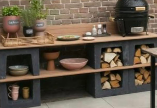 Outdoor kitchen counter with herbs, a grill, dishes, and stacked firewood set against a brick wall.