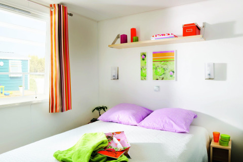 Lyst soveværelse med lilla puder, farverige dekorative elementer og sollys, på Stacaravan Confort i Frankrig.