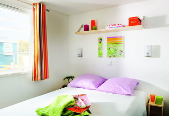 Helles Schlafzimmer mit lila Kissen, bunten Dekorationen und Sonnenlicht im Stacaravan Confort in Frankreich.