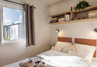 Lichte slaapkamer in Stacaravan Confort bij Flower Camping Au Bois Des Biches, Frankrijk, met raam en planken.