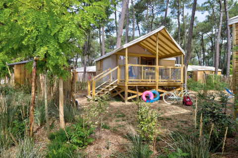 Cabane dans les arbres Cosyflower avec terrasse, vélo et bouées, entourée de verdure en forêt.
