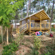 Cabane dans les arbres Cosyflower avec terrasse, vélo et bouées, entourée de verdure en forêt.