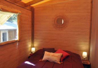 Accogliente camera in legno con letto matrimoniale, cuscini, lampade e specchio in una casa sull’albero in Francia.