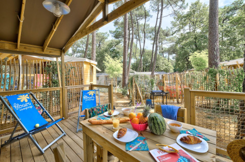 Holzterrasse der Sweetflower-Lodge im Flower Camping Au Bois Des Biches mit Frühstückstisch und Liegestühlen.