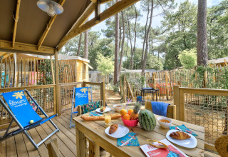 Holzterrasse der Sweetflower-Lodge im Flower Camping Au Bois Des Biches mit Frühstückstisch und Liegestühlen.