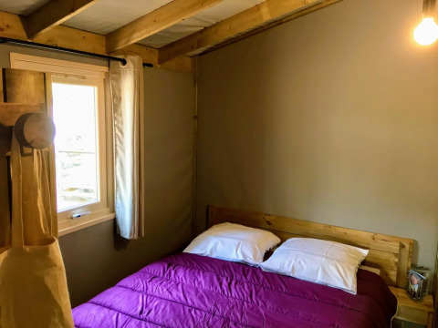 Gemütliches Schlafzimmer mit Holzbalken, Doppelbett mit lila Bettdecke und Fenster in der Sweetflower Lodge.