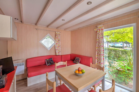 Interno di un cottage moderno con divano rosso, tavolo da pranzo e ampia finestra a Flower Camping Au Bois Des Biches, Francia.
