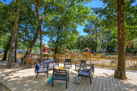 Terras van chalet met zitplaatsen onder bomen, houten omheining en zonnige kinderspeelplaats zichtbaar.