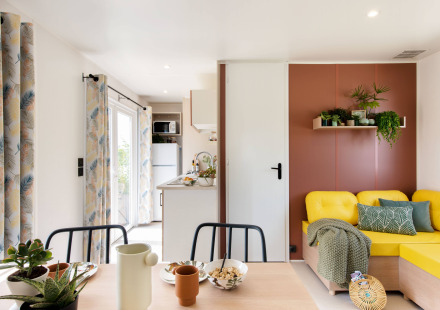 Lichte en kleurrijke chaletinterieur met gele bank, planten en eettafel op Flower Camping Au Bois Des Biches.