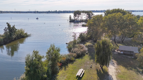 Vista di un alloggio glamping sul lago con tavolo da picnic, alberi, un riparo e riflessi di sole sull’acqua.