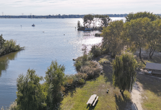 Zicht op een glampingverblijf bij het meer met picknickbank, bomen, hut en zonnelicht op het water.