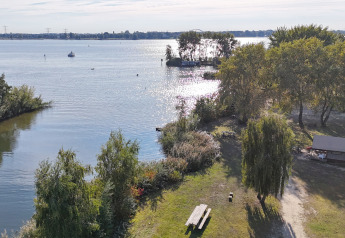 Vista de un alojamiento glamping junto al lago con mesa de picnic, árboles, refugio y sol sobre el agua.