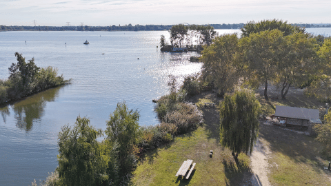 Vista de un alojamiento glamping junto al lago con mesa de picnic, árboles, refugio y sol sobre el agua.