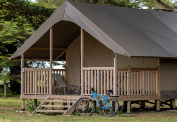 Safari telt med træterrasse, liggestole og blå cykel foran, omgivet af træer på Flower Camping Monplaisir, Frankrig.