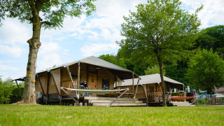 Safari tent Cottage with porch and hammock on a green campsite surrounded by lush trees, outdoors.