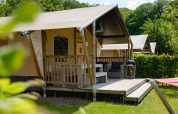 Tenda safari con veranda in legno, salotto da esterno e barbecue, immersa nel verde in una giornata soleggiata.