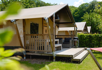 Safari tent with wooden porch, outdoor seating, and grill, surrounded by greenery and trees on a sunny day.