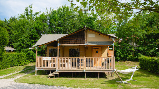 Photo of a cozy safari tent with a porch and hammock, surrounded by lush green trees and bushes.