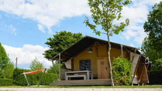 Safari tent Nomad on a campsite, featuring a wooden deck, bench, hammock, and lush greenery under blue sky.
