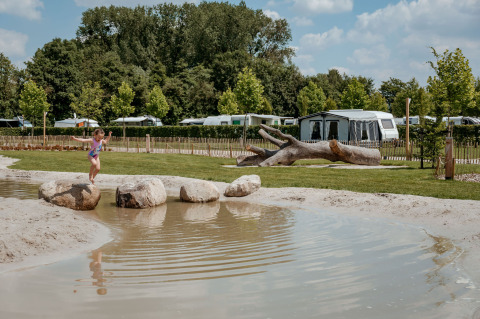 Een kind springt op stenen bij een vijver bij Limburg Lodge, Camping 't Geuldal, Nederland, met tenten op de achtergrond.