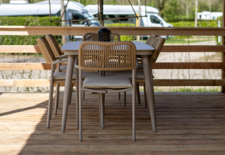 Buiteneethoek bij een lodge met houten terras, moderne tafel en stoelen, en campers op de achtergrond.