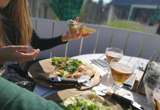 Twee personen genieten samen van pizza en bier aan een buitentafel bij een glampingverblijf in de zon.