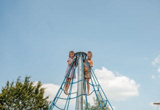 Two children climb a blue rope structure under a blue sky at Campsite 't Geuldal in Limburg, Netherlands.